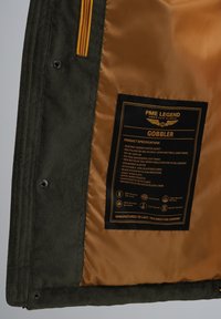 Chaqueta puffer con capucha en tela de corduroy verde oliva y forro interior dorado. Incluye cremalleras, bolsillos y una etiqueta de especificación del producto.