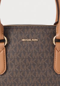 Καφέ τσάντα χειρός με μοτίβο, κατασκευασμένη από υλικό συνθετικής υφής, με χρυσές μεταλλικές λεπτομέρειες και καφέ δερμάτινα στοιχεία με λεπτομέρεια λογότυπου.