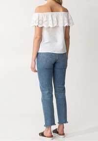 Top bianco off-the-shoulder con dettagli di pizzo ondulato, abbinato a jeans in denim blu a vita alta con orli grezzi.
