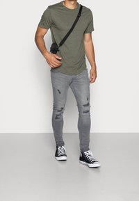 Camiseta verde oliva, vaqueros ajustados grises con desgaste, zapatillas altas en blanco y negro, llevando un pequeño bolso cruzado negro.