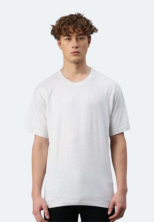 Junger Mann mit lockigem Haar, der ein einfaches hellgraues T-Shirt mit kurzen Ärmeln trägt und vor einem neutralen Hintergrund steht.