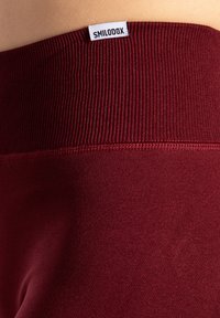 Leggings atletici color bordeaux con cintura a coste e una piccola etichetta bianca con la scritta "SMILODOX." Il tessuto ha una superficie liscia.