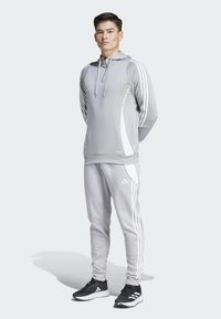 Hoodie de sport gris clair avec des accents blancs, doté d'une capuche avec cordon. Complété par un pantalon de survêtement gris et des baskets noires avec des rayures blanches.