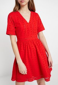 Robe rouge avec un décolleté en V, des manches courtes et un tissu texturé aux motifs élaborés. La taille est soulignée par une jupe froncée.