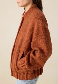 Veste en polaire texturée couleur rouille avec un col arrondi, des poches latérales et un ourlet côtelé. Coupe décontractée avec une sensation douce et confortable.