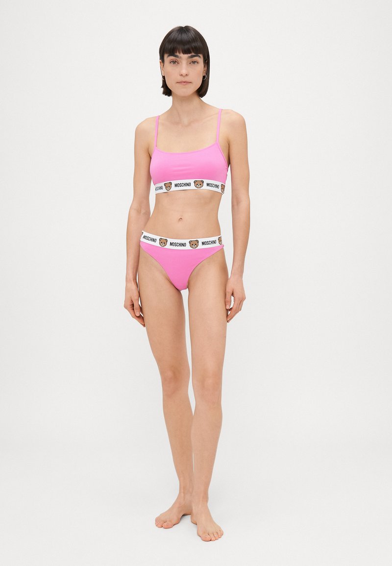 Rozā bikini komplekts ar pieguļošu braleti ar plānām siksnām un atbilstošām zemāka piegriezuma biksītēm, abiem ar Moschino lāča logo jostu.