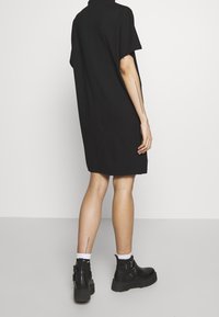 Musta, rento-mallinen mekko, jossa on lyhyet hihat ja korkea kaulus. Materiaali näyttää sileältä ja se on yhdistetty massiivisiin mustiin nilkkasaappaisiin ja valkoisiin sukkiin.