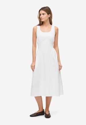 Kvinde stående iført en hvid ærmeløs midikjole med samlinger i siderne og sort vævet flad sko, kigger let mod venstre.
