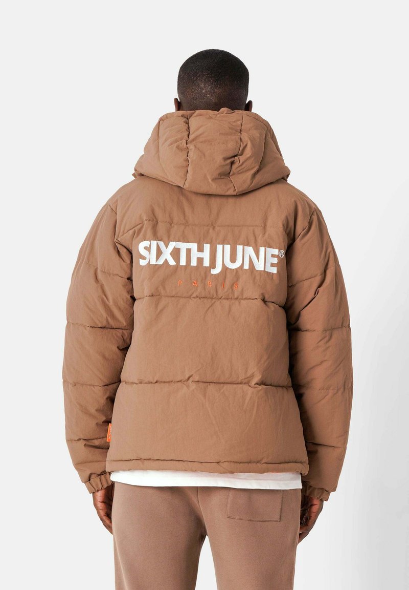 Persoon in een bruine pufferjack met "SIXTH JUNE PARIS" in het wit en oranje op de achterkant, staand tegen een eenvoudige achtergrond.
