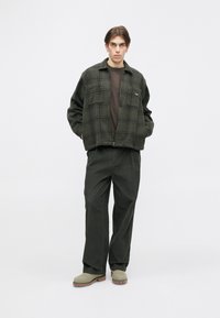 Zelena karirana volnena jakna preko rjave srajce, v kombinaciji z ohlapnimi temnimi hlačami in sivimi klobuki, stoji na enostavni podlagi.