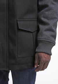 Schwarze Wollmischung Jacke mit strukturierter Oberfläche, ausgestattet mit einer Klapptasche und gerippten Bündchen. Der Ärmel zeigt eine Hand, die nahe der Tasche ruht.