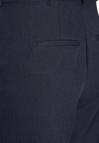 Närbild av marinblå skräddarsydda byxor som visar en sidfick med kantband och herringbone-tygstruktur.