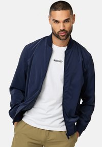 Marine bomberjakke lavet af glat stof med ribbede manchetter og kant. Bæres over en hvid T-shirt med sort tekst. Synlig lynlåsdetaile.