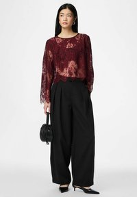 Sieviete valkā caurspīdīgu bordo krāsas mežģīņu topu ar garām piedurknēm, melnas platas bikses, melnus augstpapēžu kurpes ar smailiem purngaliem un tur nelielu melnu rokassomu.