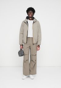 Doudoune beige clair avec zip noir et jaune, t-shirt blanc, pantalon large kaki et baskets blanches ; tenant un petit sac gris.