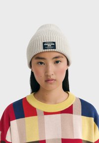 Ung kvinde iført en beigefarvet strikhue med et sort mærke, der står "THINKING MU", og en farverig ternet sweater foran en ensfarvet baggrund.