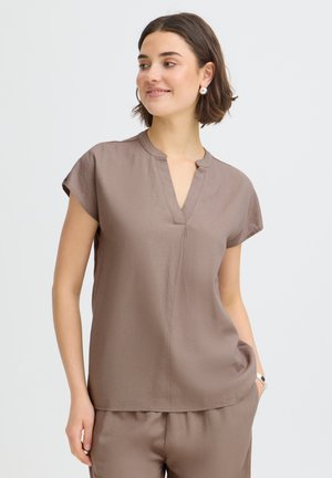 Žena s krátkymi hnedými vlasmi v béžovom krátkom rukáve s výstrihom do V a zodpovedajúcich nohaviciach, usmieva sa a pozerá do boku.