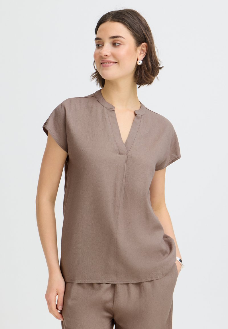 Femme aux cheveux bruns courts portant un haut taupe à manches courtes et encolure en V, ainsi qu'un pantalon assorti, souriante et regardant sur le côté.