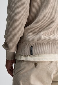 Beige gerippter Strickpullover mit strukturierter Oberfläche, ausgestattet mit einem sichtbaren Etikett am unteren Saum, getragen über einem hellen Shirt und Hose.
