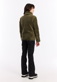 Dívka stojící zády, oblečená v olivově zelené fleecové bundě, černých kalhotách s hvězdnými vzory a černých botách s bílými podrážkami na bílém pozadí.