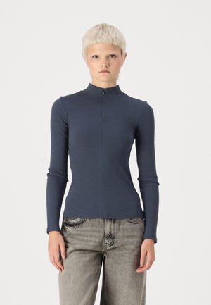 Navy ribbed long-sleeve top with a high neck and front zip, paired with faded gray wide-leg jeans. Smooth fabric texture.