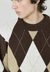 Pull en argyle marron, beige et blanc. Fait d'un matériau doux avec un col rond et des coutures blanches le long des formes en losange.