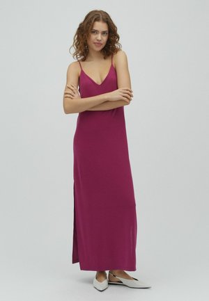 Kvinde med krøllet hår iført en ærmeløs, dyb magenta maxi-kjole og hvide spidse flade sko, stående med armene krydsede på en ensfarvet baggrund.