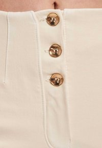 Nærbilde av beige stoffbukser med tre blanke gullknapper festet vertikalt langs området for glidelåsen foran.