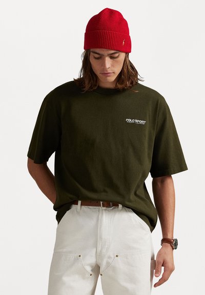 Fiatal személy piros kötött sapkát, sötétzöld Polo Sport Ralph Lauren pólót, fehér nadrágot barna övvel és karórát visel, és lefelé néz.