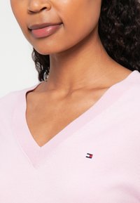 Maglione rosa chiaro a scollo a V realizzato in tessuto a maglia, con un piccolo logo ricamato sull'area sinistra del petto. Texture morbida, design casual.