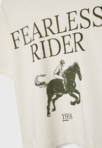 Cremefarvet bomulds t-shirt med grafisk tryk af en rytter på en hest, med teksten "FEARLESS RIDER" og året "1918."