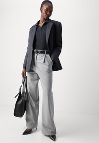 Pantalons gris sur mesure avec taille haute et plis à l'avant, associés à un polo noir et un blazer rayé. Les accessoires comprennent un sac à main noir.