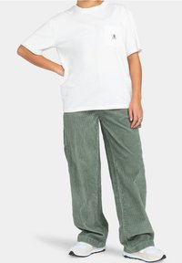 T-shirt en coton blanc avec une poche poitrine et un logo, associé à un pantalon en velours côtelé vert olive, ample et texturé. Des baskets blanches complètent la tenue.
