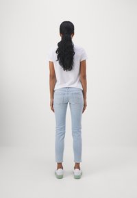 Camiseta blanca de manga corta, jeans ajustados azul claro y zapatillas blancas con detalles verdes. Costuras visibles en los pantalones y en los bolsillos traseros.