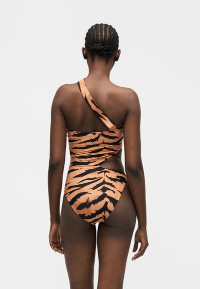 Γυναίκα με πλεξούδες στα μαλλιά που φοράει ολόσωμο μαγιό με μία τιράντα σε μοτίβο τίγρης και άνοιγμα, εμφανιζόμενη από πίσω σε απλό γκρι φόντο.