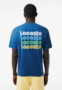 Camiseta de manga corta azul con el texto apilado "Lacoste" en tonos blancos, verdes y amarillos en la parte posterior. Hecha de tela de algodón.