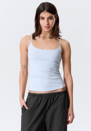 Sieviete ar plecu garumu tumšiem matiem, tērpusies gaiši zilā bezroku topā un melnas biksēs, stāvot ar vienu roku kabatā pret vienkrāsainu fonu.