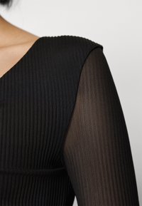 Černý top s žebrovanou texturou na těle a dlouhými rukávy z průhledné síťoviny. Výstřih do V zvýrazňuje dekolt.
