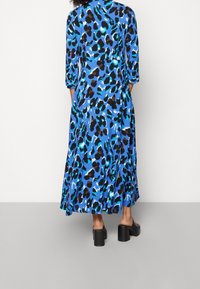 Langes blaues Kleid mit schwarzem und türkisfarbenem Leopardenmuster. Mit Rundhalsausschnitt und dreiviertel Ärmeln, fließendem Rock. Schwarze klobige Absätze.