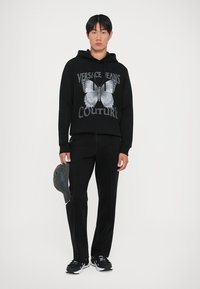 Hanorac negru cu un design grafic de fluture, blugi negri și o șapcă gri ținută într-o mână. Încălțămintea are accente negre și albe.