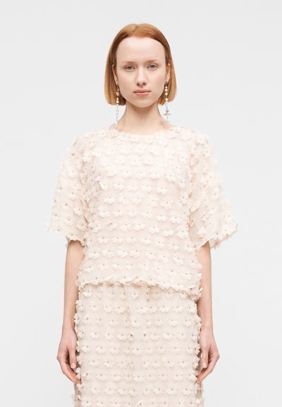 Ροδακινί τοπ με 3D εφαρμογές λουλουδιών, διαφανή υφή, άνετη εφαρμογή και φαρδιά μανίκια, σε συνδυασμό με φούστα με λουλουδάτο σχέδιο που ταιριάζει.