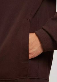 Brązowa bluzka z materiału o gładkiej teksturze posiada boczną kieszeń i ściągacze przy mankietach, prezentując dopasowany krój i minimalne detale szwów.