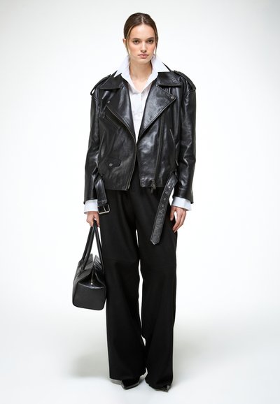 Žena u crnoj kožnoj jakni, bijeloj košulji s ovratnikom, crnim širokim hlačama i držeći crnu torbu, stoji na bijeloj pozadini.