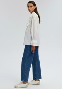 Camicetta bianca con dettagli arruffati e inserti in pizzo, abbinata a pantaloni in denim blu a vita alta e sneakers bianche con rifiniture verdi.