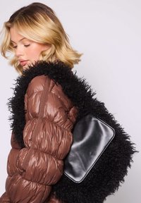 Blondynka w brązowej pikowanej kurtce, niosąca dużą czarną teksturowaną torebkę na ramieniu z gładkim czarnym panelem ozdobnym, patrząca w dół.