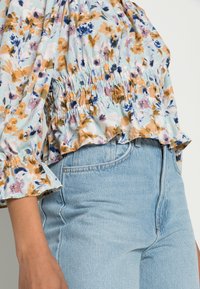 Blouse fleurie en tissu bleu clair avec smocks élastiques à la taille, présentant un mélange de fleurs orange, rose et bleues. Coupe décontractée avec des poignets volantés.