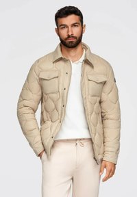 Uomo con barba che indossa una giacca trapuntata beige, una camicia bianca e pantaloni beige chiaro, in piedi di fronte a uno sfondo chiaro e uniforme.