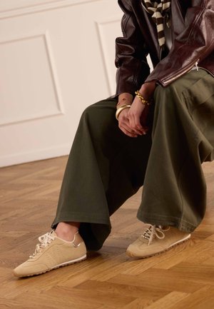 Persoon zittend, gekleed in olijfgroene broek met wijde pijpen, beige sneakers, bruin leren jasje, gestreepte sjaal en gouden armbanden, met handen gevouwen.