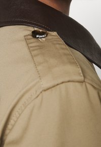 Zbliżenie na ramię beżowej kurtki z ciemnobrązowym skórzanym kołnierzem i czarnym guzikiem z napisem "PINKO".