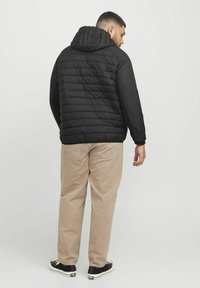 Black puffer jacket with a hood, featuring quilted sections. Worn with light beige pants and black shoes, showing a casual back view.